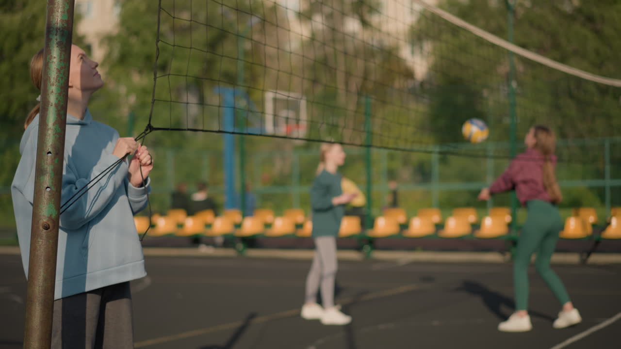 두 명의 여성이 배경에서 볼리볼을 치고 다른 사람들이 멀리서 흐릿하게, 밝은 빛으로 야외 스포츠 설정
