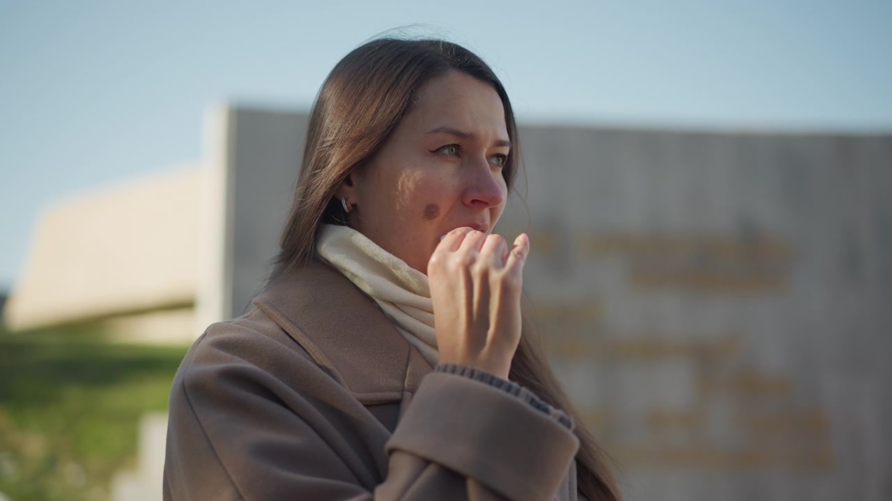 Primo piano di una donna all'aperto che si sistema la sciarpa con una mano, mentre indossa un cappotto marrone e uno scaldacollo color crema. Lo sfondo presenta una struttura in cemento sfocata sotto la luce chiara del giorno.