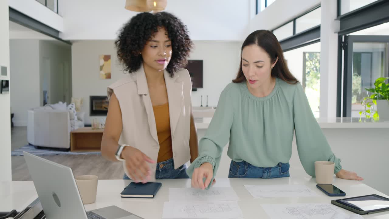 Colleghe donne di diverse etnie che osservano un piano stampato, appoggiate a un tavolo di casa, srotolano i piani e finalizzano il design.