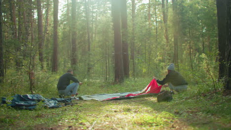 lovers prepare tent for camping in forest as man holds arched pole and lady presses fabric to ground securing it in place, sunlight creates gentle glow around her