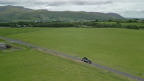 Black-car-driving-along-countryside-road-with-mountain-Blencathra-in-background-on-cloudy-summer-day