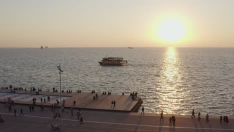 aerial - people walking by the sea in thessaloniki with a boat in the background at sunset