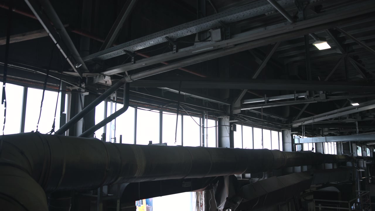Bred scene fra lav vinkel inde i et industrielt tagrum, der viser en overdimensioneret isoleret ventilationskanal, der løber hen over forgrunden under synlige stålfagværk, diagonale støtteafstivninger, kabelbakker og