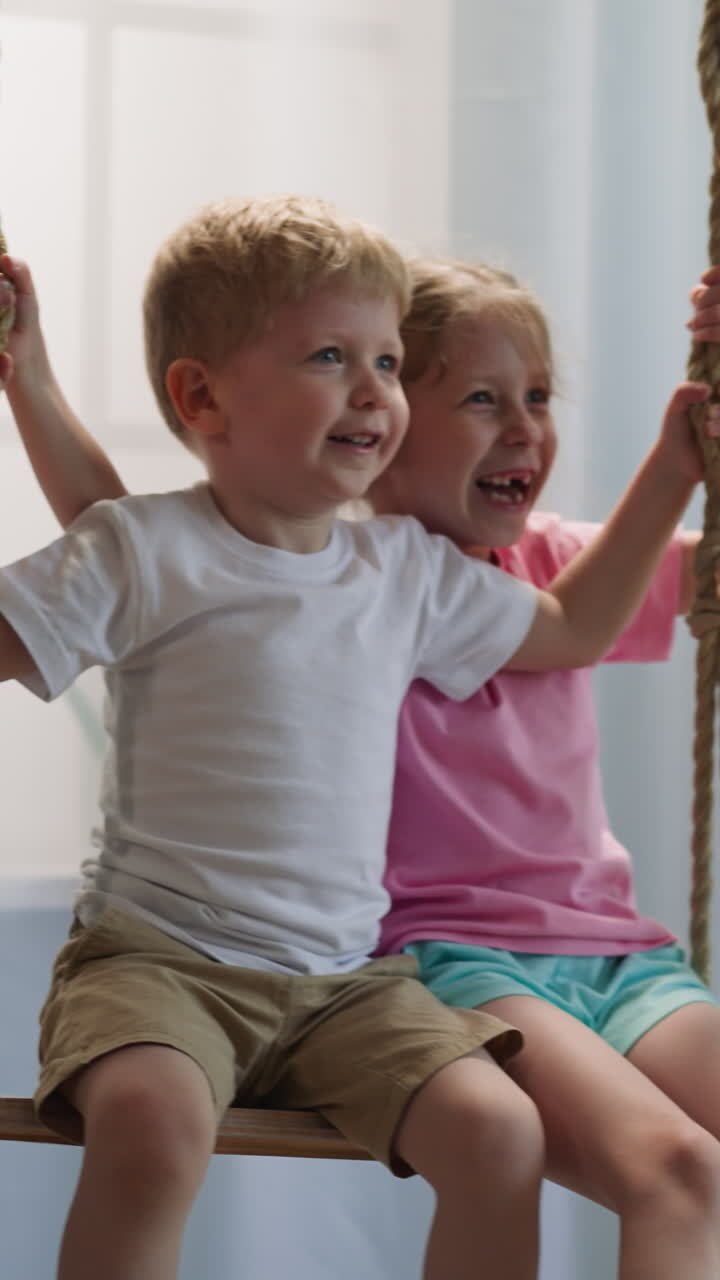 Toddler boy and preschooler girl play together in light room with window and bookcase. Friendly children siblings chat cheerfully riding on swing at home