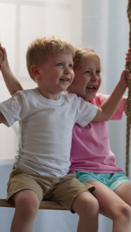 toddler boy and preschooler girl play together in light room with window and bookcase. friendly children siblings chat cheerfully riding on swing at home