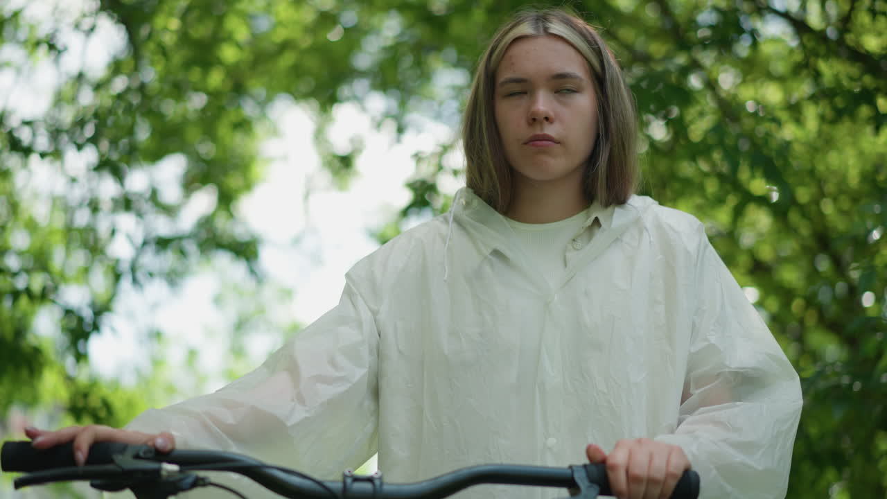 молодая женщина в полупрозрачном плаще останавливается, снимая руку с руля велосипеда, в то время как всматривается в расстояние, фон наполнен размытой зеленой листьями