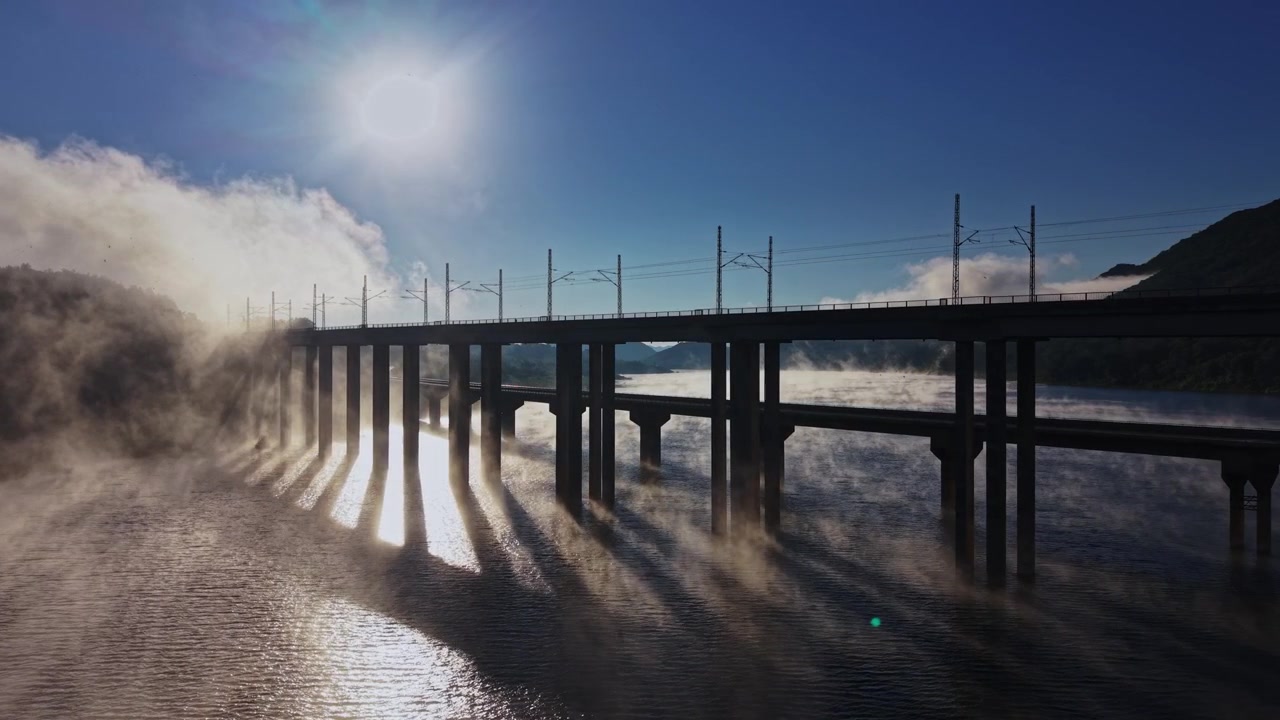 太陽が輝く空の下、川の橋にかかる朝霧