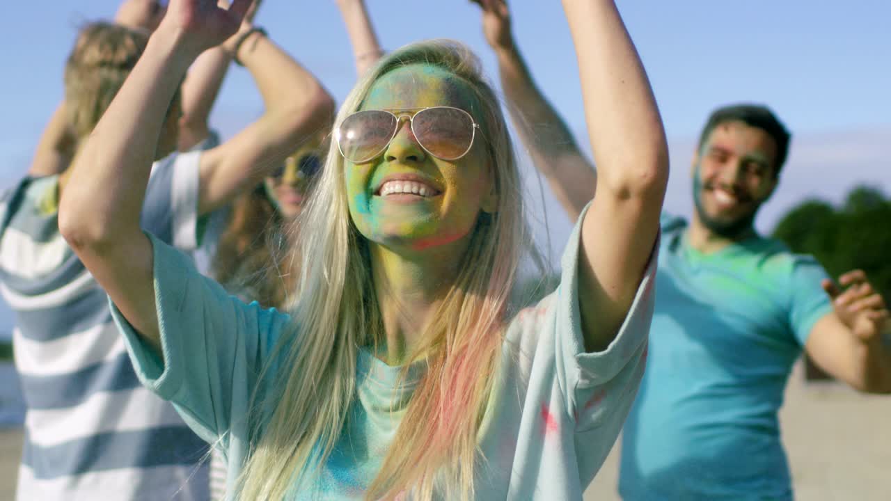 Close-up Portrait of a Beautiful Blonde Girl Dances in Celebration of Holi Festival With Her Friends. Her Face and Clothes are Covered with Colorful Powder. People Dancing in the Background.