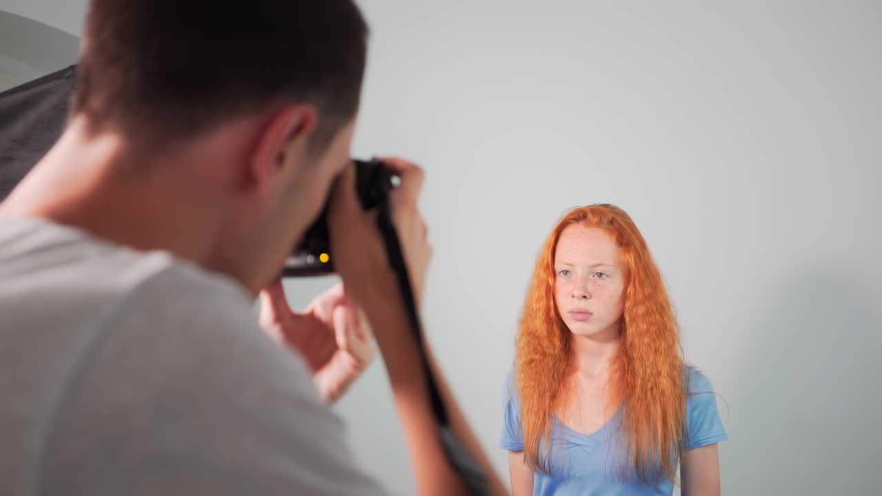 Young professional photographer taking photos of a red haired girl in a studio with white background. Rear view. Girl with lots of freckles on the face. Slow motion