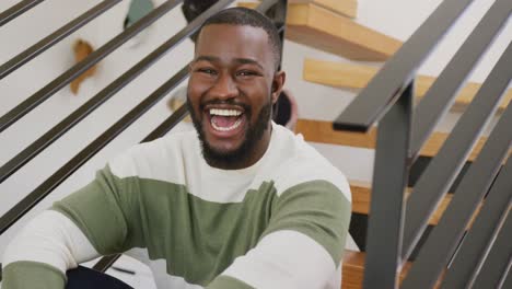 video portrait of happy african american man laughing to camera, sitting on stairs at home