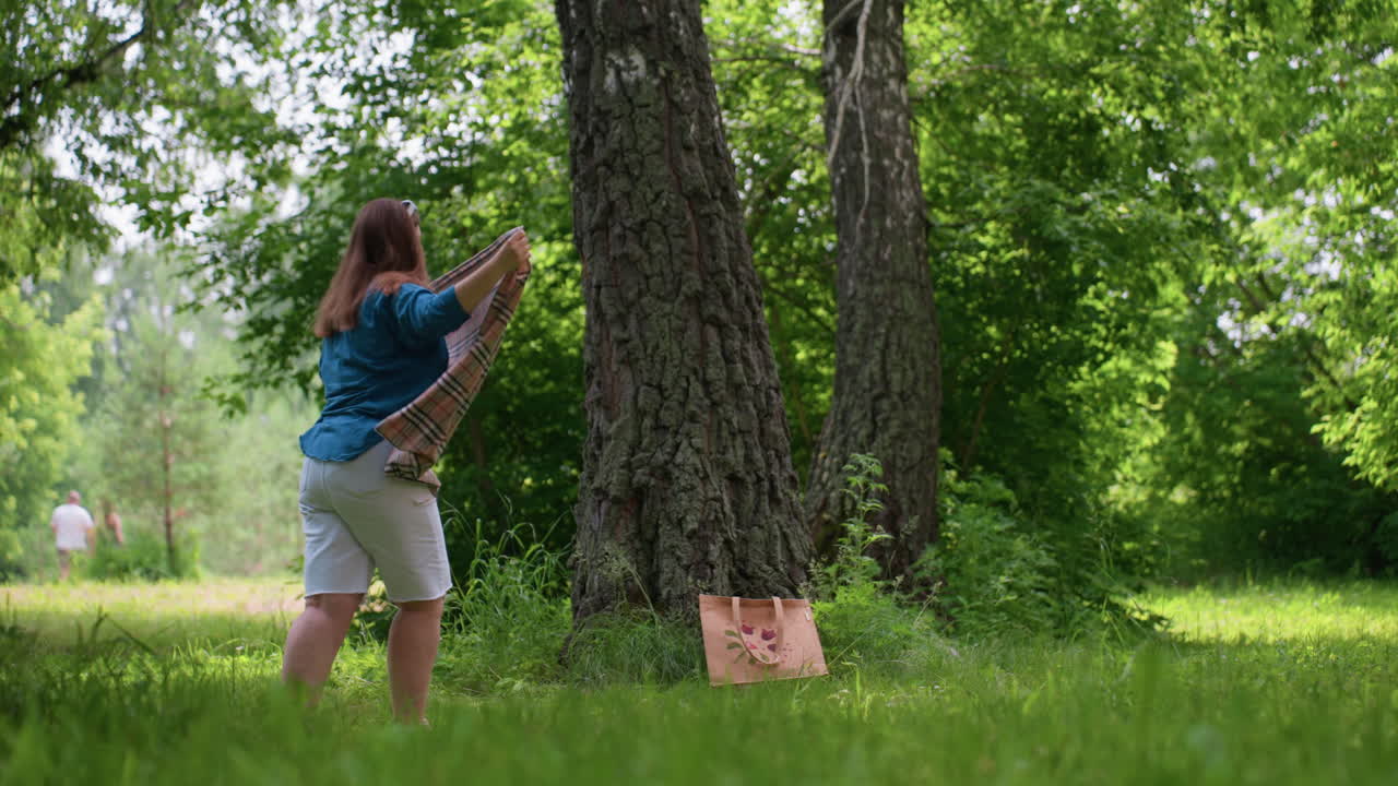 College student folds blanket carefully in green park surrounded by trees, soft sunlight, and gentle breeze, with blurred figures of people walking in distance