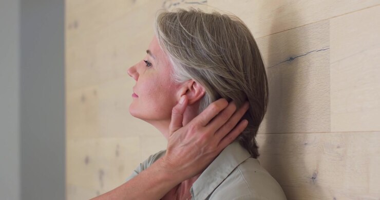 Donna anziana appoggiata a una parete di legno, alzando la mano per accarezzarsi i capelli e rilassare il collo, in camicetta.