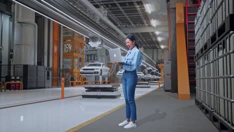 vista lateral completa del cuerpo de una trabajadora profesional asiática de pie con su portátil en una fábrica de automóviles ev, instalación industrial de alta tecnología, levanta el puño con una meta gritando después de escribir en la portátil