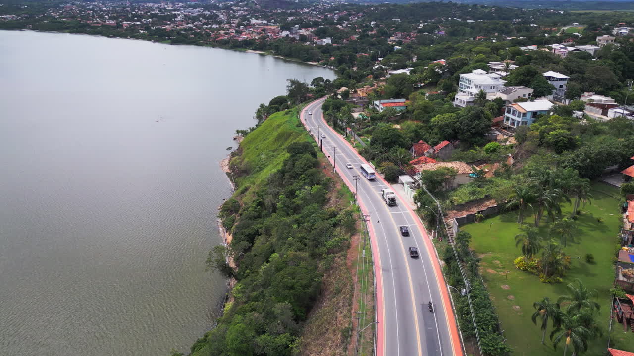 A tracking drone follows vehicles along a road beside a lagoon in Saquarema, home to a World Surf League venue. The lagoon on one side houses on the other, nearby Mirante do Morro da Cruz.