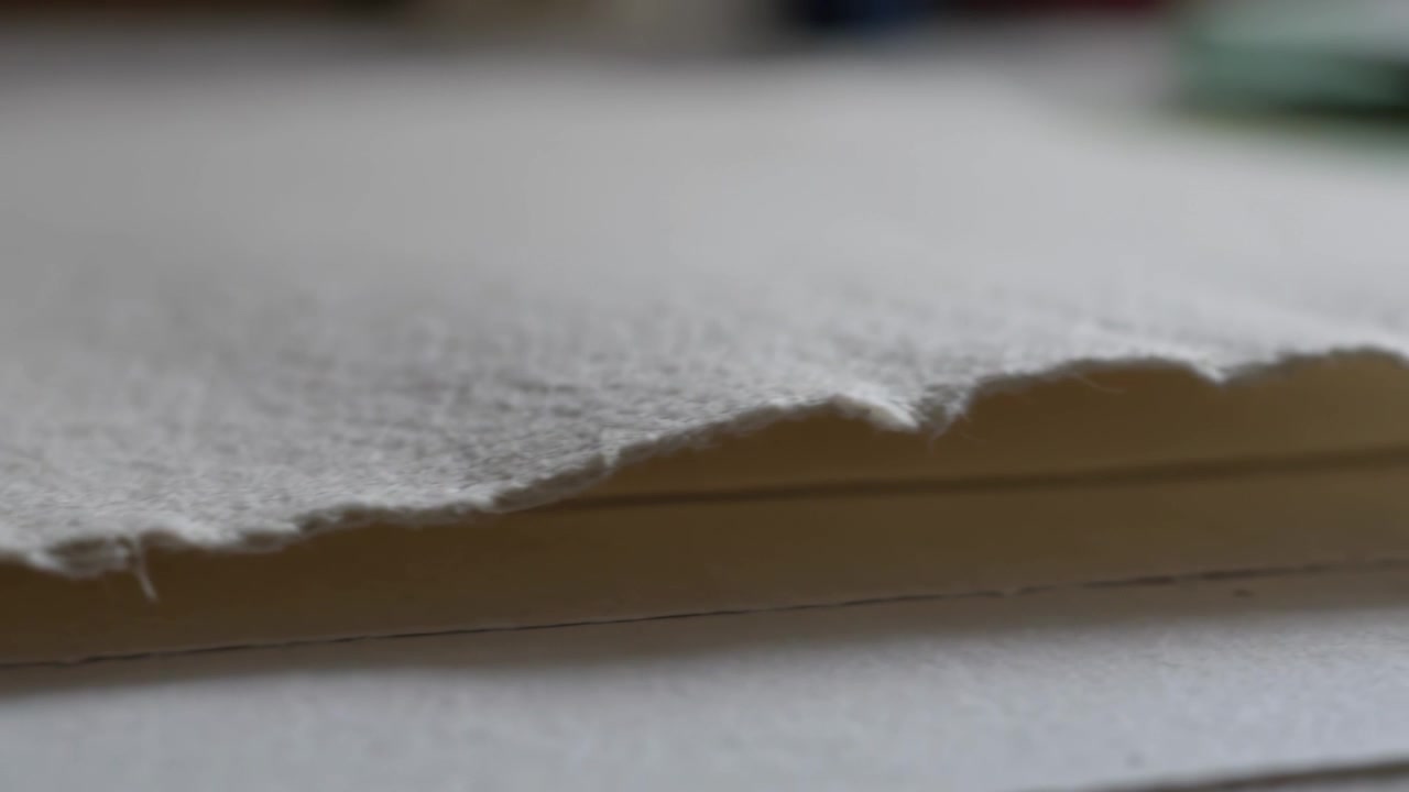 Pulling focus, camera showing deckled-paper stack on desk showing thin line, green blur and texture