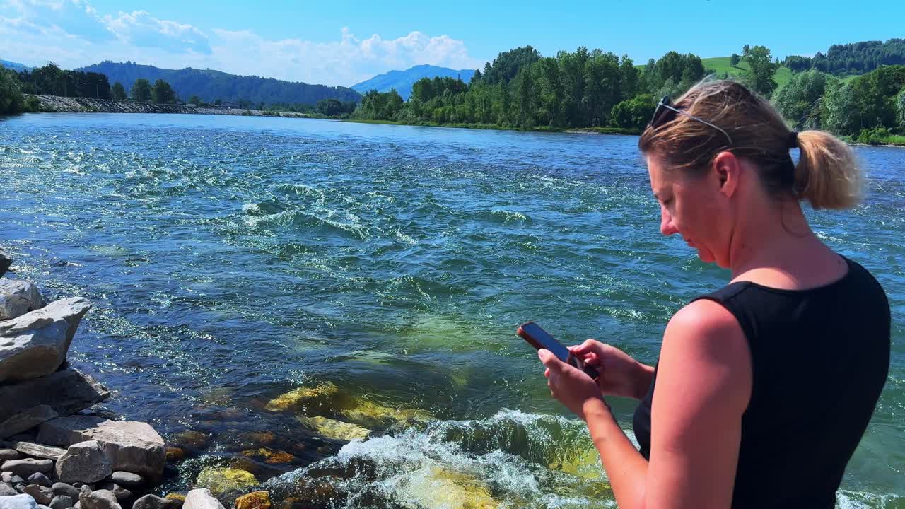 Kobieta pisząca SMS-a nad rzeką, oddająca spokój i witalność natury, z płynącą rzeką w tle pod czystym, błękitnym niebem.