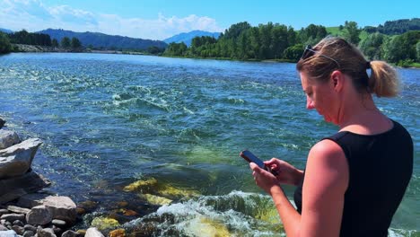 woman engaged in texting by the riverside, capturing nature's serenity and vibrancy with a flowing river in the background under clear blue skies