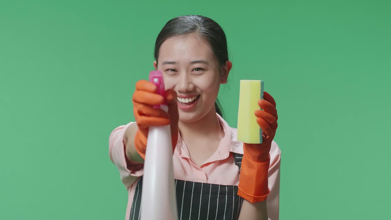 초록색 스크린 스튜디오에 서 있는 동안 주방 스폰지를 들고 스프레이를 사용하는 아프론을 입은 미소 짓는 아시아 여성 집사.