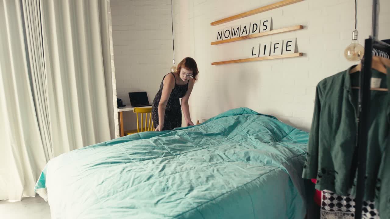 여름 드레스와 안경을 입은 독신 여성이 침실에서 침대를 짓습니다.