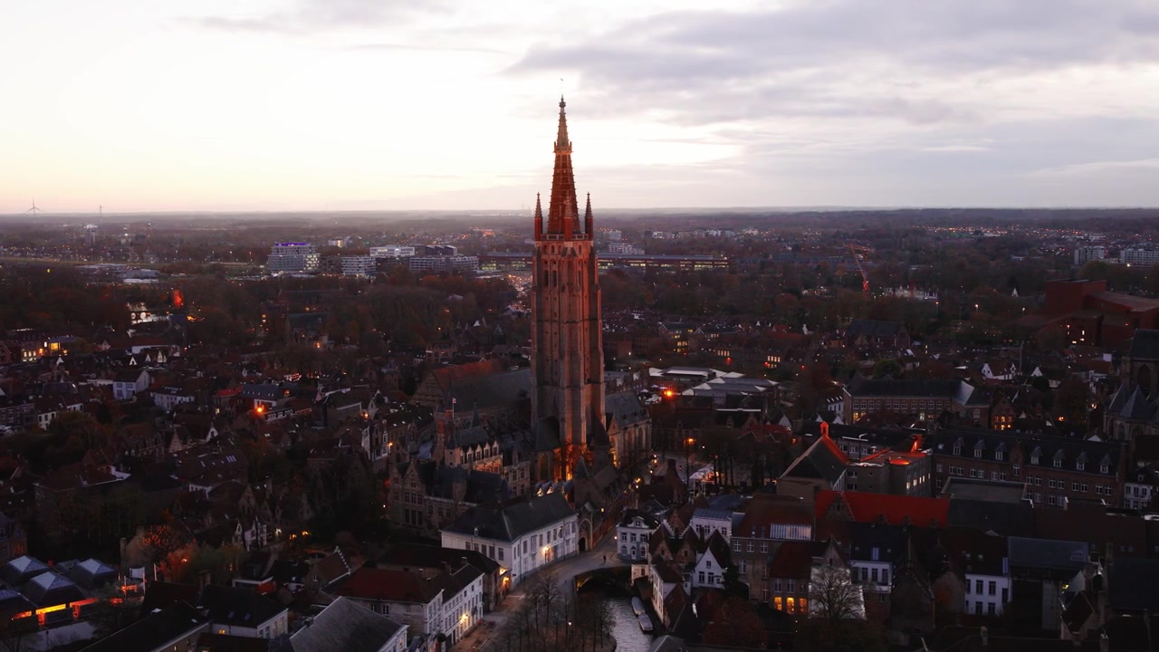 Filmagem de drone da Igreja de Nossa Senhora em Bruges ao entardecer, mostrando arquitetura iluminada e uma bela vista panorâmica da cidade histórica.