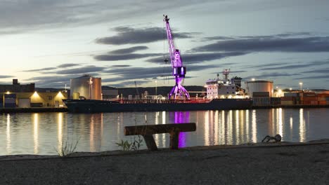 Motion-timelapse-of-illuminated-pink-crane-with-cargo-ship-by-harbour