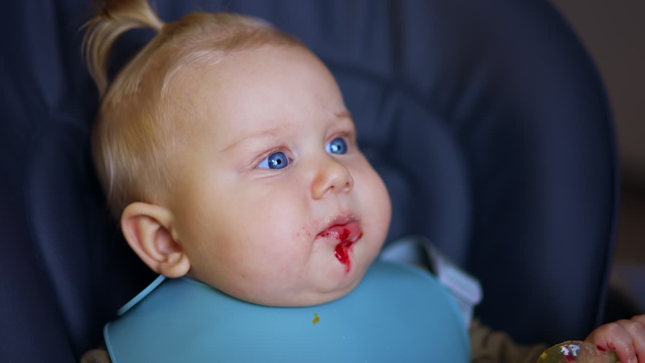Face of a cute chubby Caucasian baby with food on his chin. Close up. Kid takes a spoon from his mom and pulls it to his mouth.