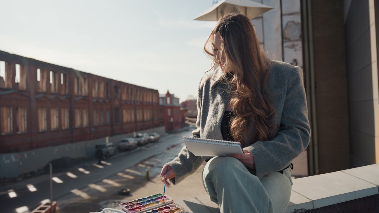 Bella giovane donna che dipinge su un tetto, sfondo industriale illuminato dal sole con edifici in mattoni, tavolozza e pennelli accanto a lei, posa rilassata, caldo bagliore dorato, espressione pensierosa, skyline urbano.