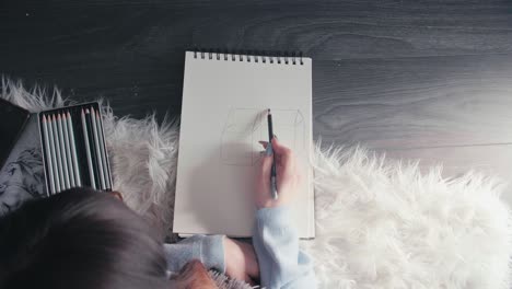 sketching on paper notebook with charcoal pencil, top down of young artist student sitting on white hair rug drawing on white paper developing new artistic creative skill