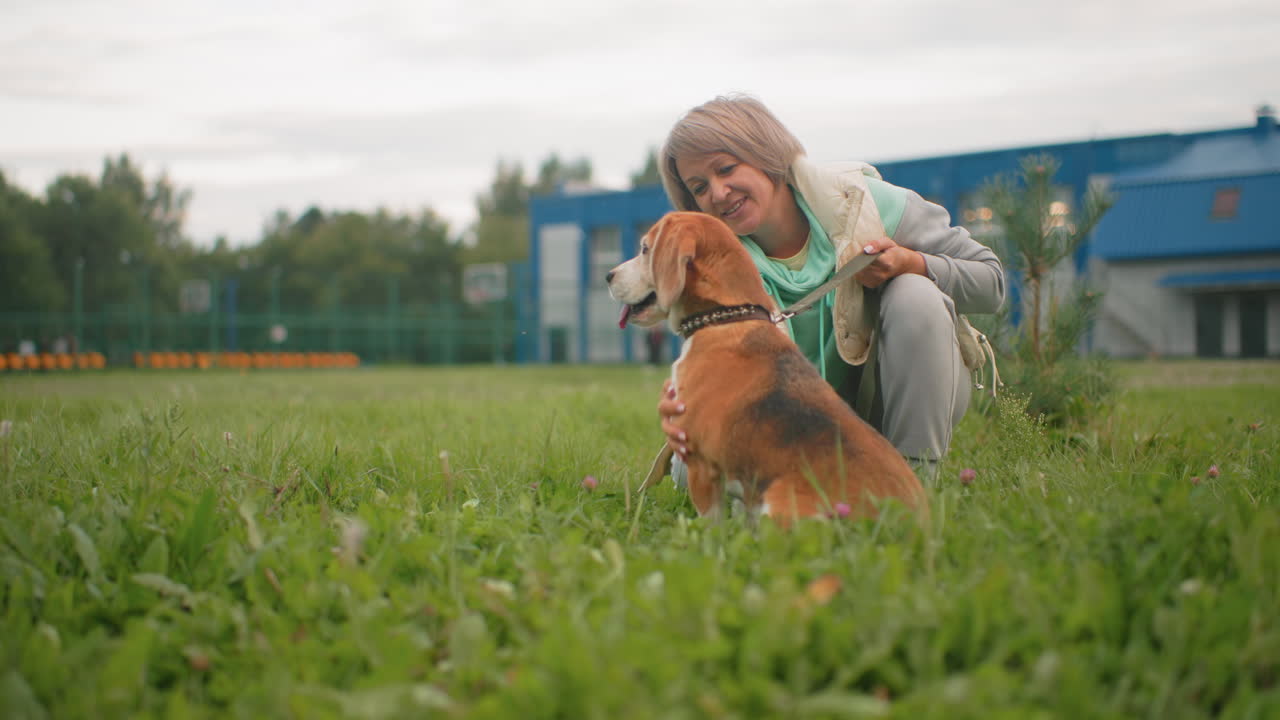 Happy dog owner in light jacket and mint green hoodie lovingly petting her puppy while kneeling on grassy field near modern buildings on cloudy day, creating joyful bond and playful outdoor moment