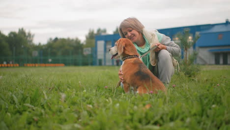 happy dog owner in light jacket and mint green hoodie lovingly petting her puppy while kneeling on grassy field near modern buildings on cloudy day, creating joyful bond and playful outdoor moment
