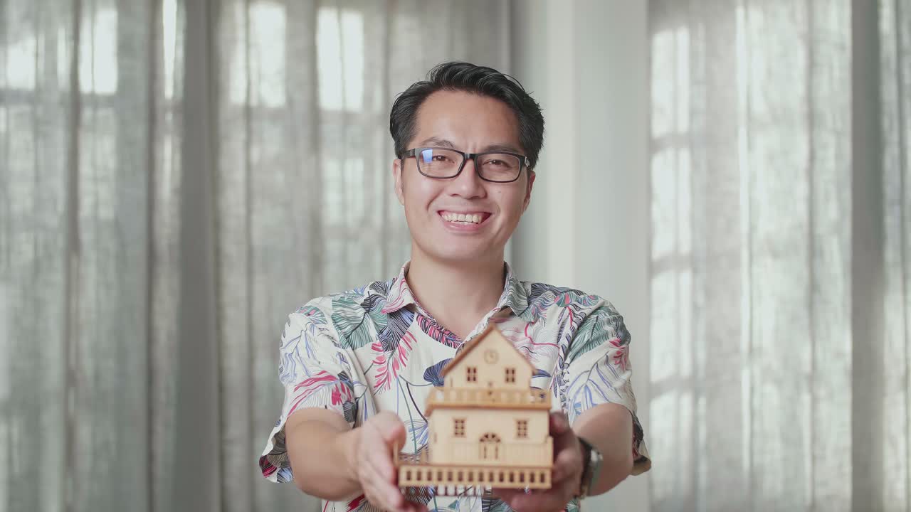 Tampilan Dekat Pria Asia dengan Kotak Kardus Tersenyum dan Menunjukkan Model Rumah ke Kamera di Rumah Baru