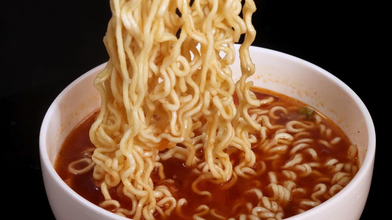 A pair of chopsticks lifts steaming instant noodles from a white bowl filled with broth, under bright studio lighting against a black background. Smooth camera movement, close-up view