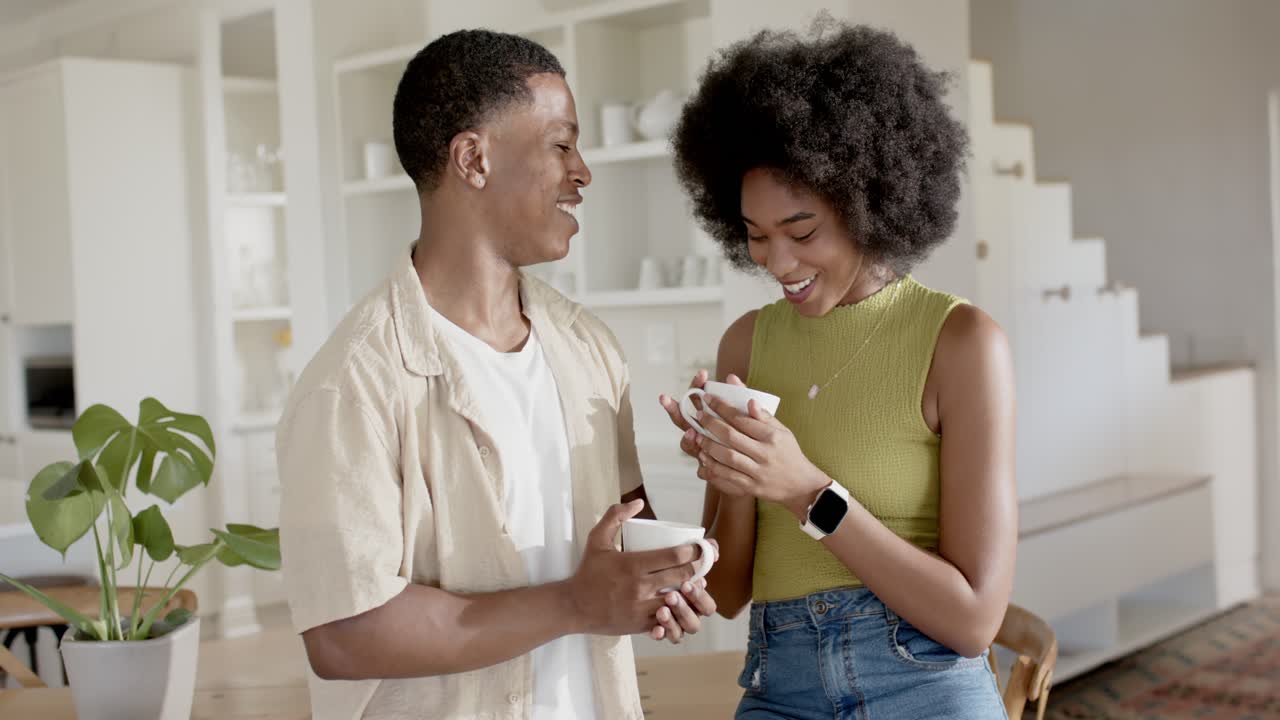 Coppia afroamericana che condivide il caffè del mattino, con tazze bianche in mano, sorridente, appoggiata in cucina.