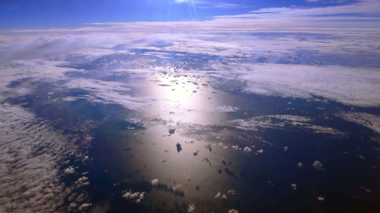 A high altitude aerial view over clouds on a sunny day, taken from the cabin of a passenger aircraft. The camera is stationary, tilted down looking out the port side window at the ocean and clouds.