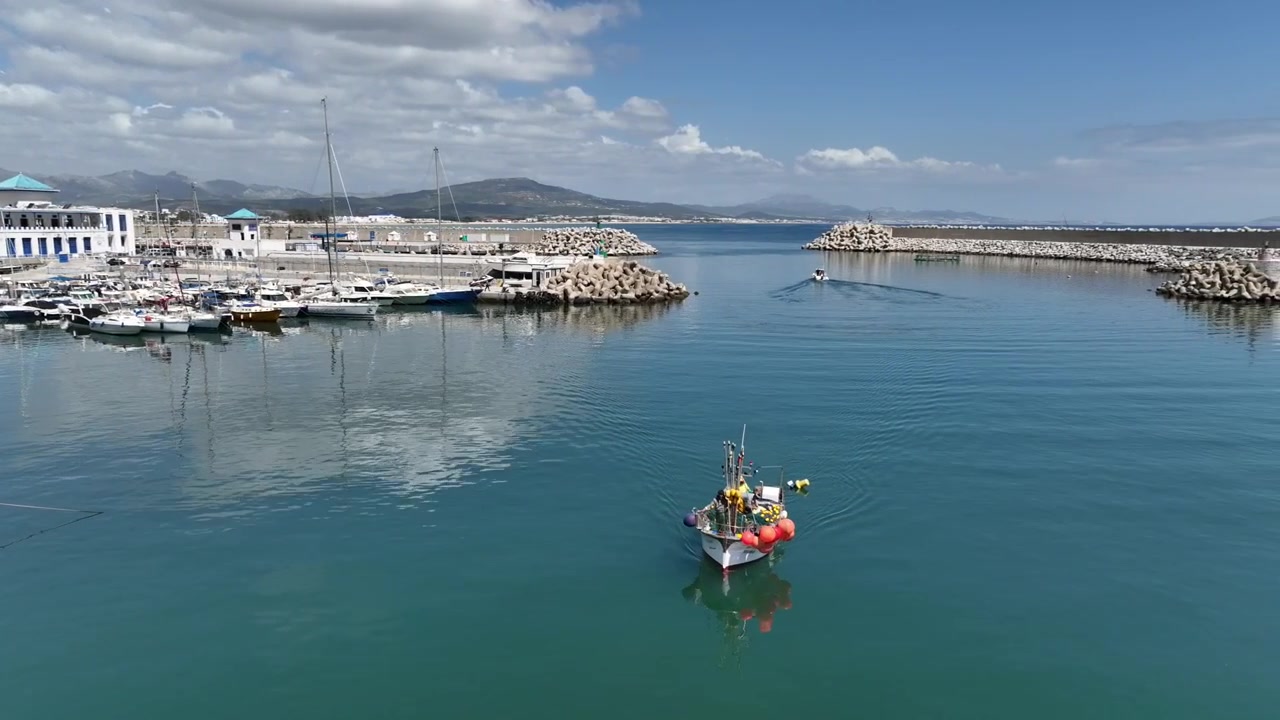 Ampia vista aerea di un porto mediterraneo attivo nel Marocco settentrionale, che mostra piccole imbarcazioni da pesca e da diporto, strutture portuali, banchine protette e una città costiera con montagne sullo sfondo.