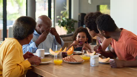 Foto-Familiar-Con-Abuelos,-Padres-Y-Nieta-Haciendo-Muecas-Durante-El-Desayuno-Alrededor-De-La-Mesa-En-Casa.