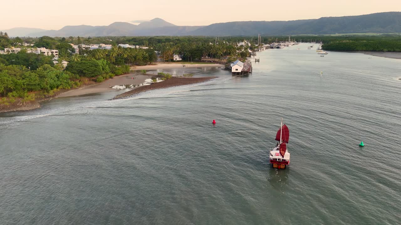 Drone captures sailboat navigating towards marina at sunset, surrounded by lush greenery and calm waters