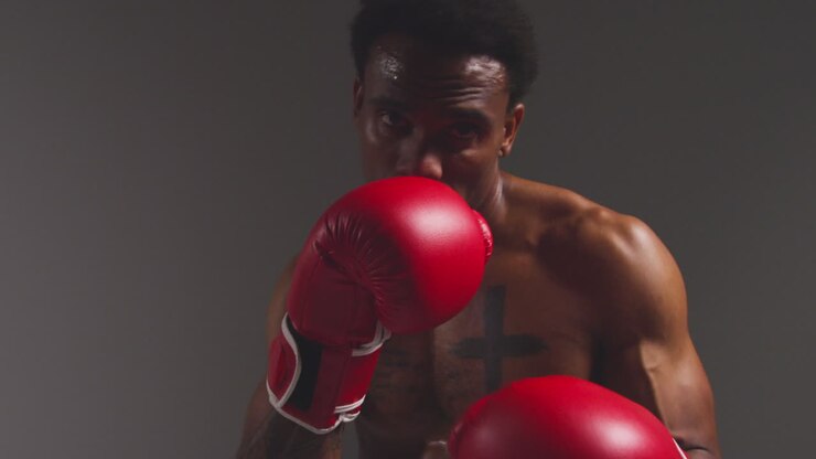 Close Up Studio POV Shot Of Tattooed Male Boxer Putting Up Gloves In Boxing Match Against Grey Background
