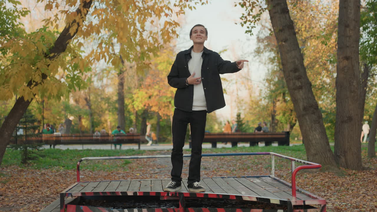 Young man standing on platform in autumn park, expressive gestures rehearsing marriage vows, golden leaves carpet ground, wooden bench and trees background, candid portrait of hopeful preparation