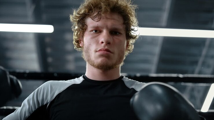 Angry kickboxer looking at camera at gym. Sportsman checking boxing gloves