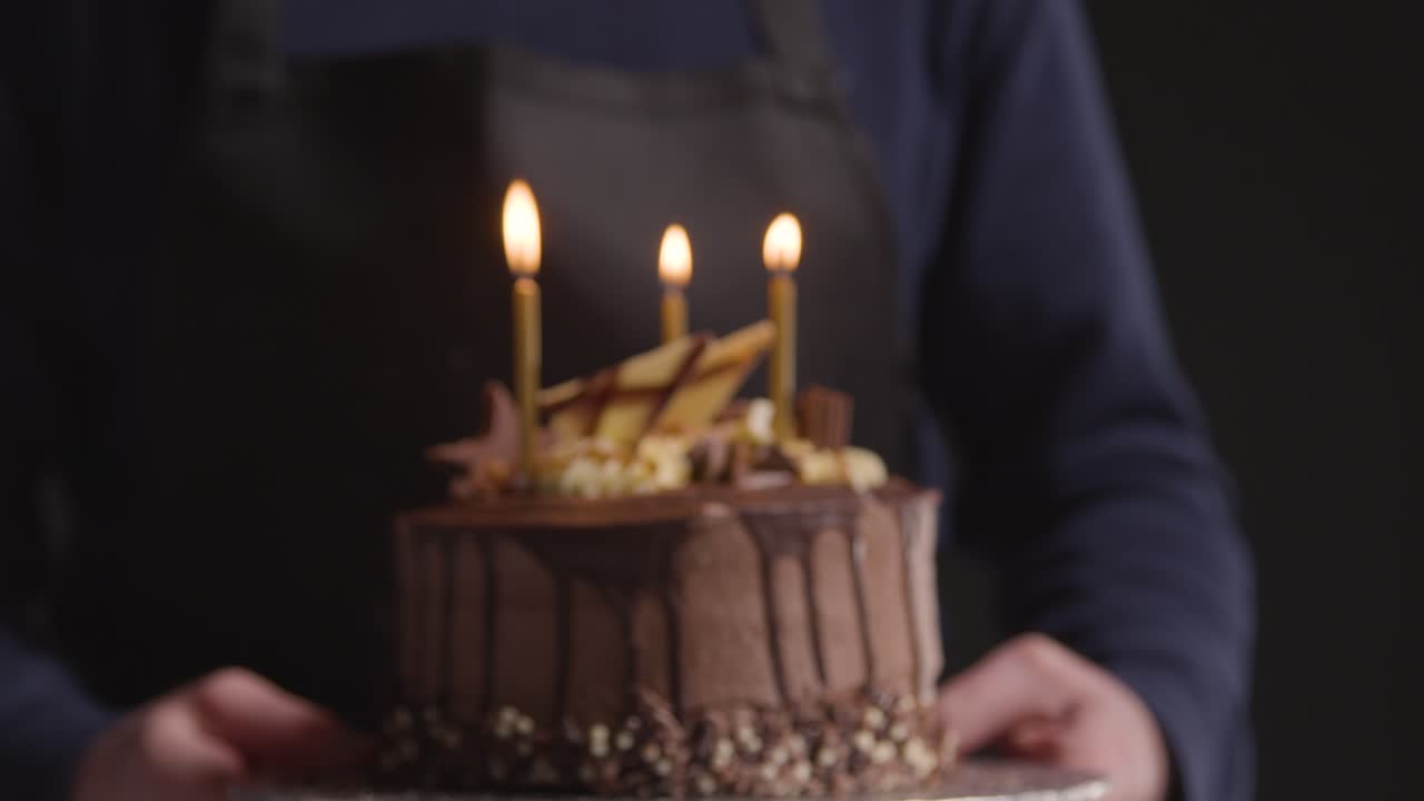 foto de estudio de una persona que lleva un pastel de cumpleaños de chocolate decorado con velas encendidas sobre fondo negro 1