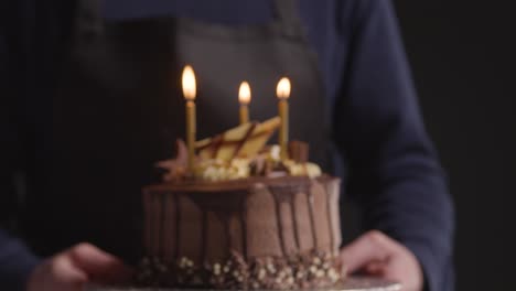 foto de estudio de una persona que lleva un pastel de cumpleaños de chocolate decorado con velas encendidas sobre fondo negro 1