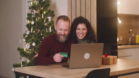 pareja comprando en línea con una tarjeta de crédito usando una computadora portátil sentada en una mesa cerca de un regalo en una habitación decorada con un árbol de navidad