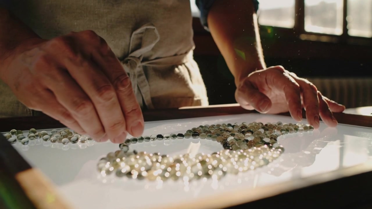 Entering craftsperson hands arranging translucent beads on backlit tray at workbench, refining loop