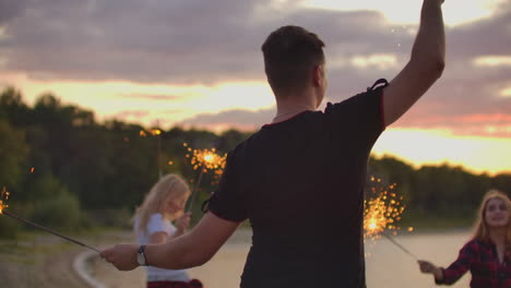 The-young-man-in-black-t-shirt-is-dancing-with-big-bengal-lights-on-the-sand-beach-with-his-friends.-This-is-crazy-summer-evening-on-the-open-air-party.