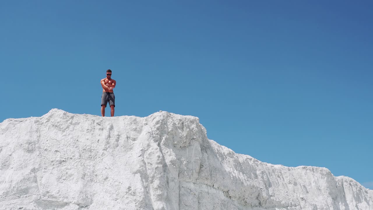 A strong man with glasses demonstrates his muscular body. Bodybuilding on the street on a white mountain against the blue sky
