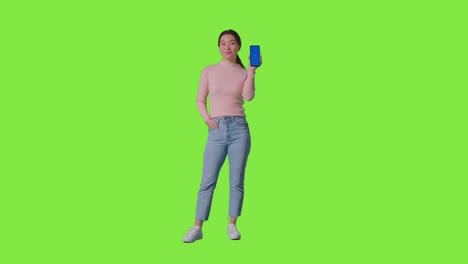 full length studio portrait of smiling woman holding blue screen mobile phone towards camera against green screen 2