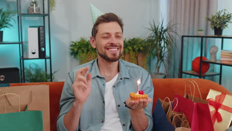 Happy-young-adult-man-celebrating-birthday-party,-makes-wish-blowing-burning-candle-on-small-cupcake