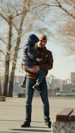 padre llevando a su hijo mientras está de pie cerca de un banco en un camino pavimentado, con un paisaje urbano en el fondo, el niño está vestido con una chaqueta hinchada