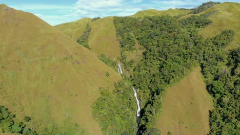 Luftaufnahme,-Annäherung-An-Einen-Wasserfall-Zwischen-Den-Bergen-Mit-Leuchtend-Grüner-Vegetation-Und-Blauem-Himmel-Im-Hintergrund